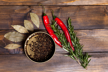 Red chili pepper and rosemary sprigs with laurel leaves and pepper black peas in a jar on wooden background. Top view of the workplace.の写真素材