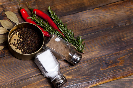 Red chili pepper and rosemary sprigs with laurel leaves and pepper black peas in a jar, shaker cans with salt and pepper on wooden background. Top view of the workplace.の写真素材