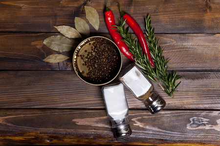 Red chili pepper and rosemary sprigs with laurel leaves and pepper black peas in a jar, shaker cans with salt and pepper on wooden background. Top view of the workplace.の写真素材