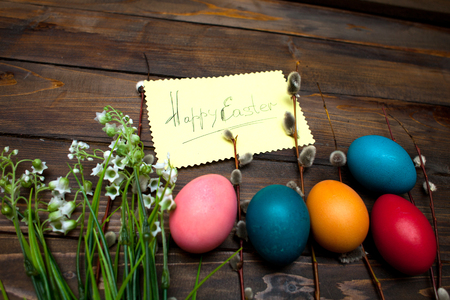 Green lily of the valley and the beautiful willow branches with bright colored Easter eggs and yellow piece of paper and the words Happy easter on wooden background.の写真素材