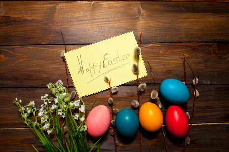 Green lily of the valley and the beautiful willow branches with bright colored Easter eggs and yellow piece of paper and the words Happy easter on wooden background.の写真素材