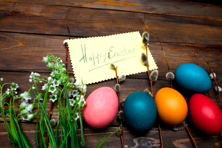 Green lily of the valley and the beautiful willow branches with bright colored Easter eggs and yellow piece of paper and the words Happy easter on wooden background.の写真素材