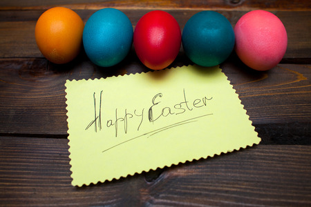 Closed, painted in different bright colors chicken eggs for the holiday Easter on natural wooden background with sheet of paper and the inscription Happy Easter.の写真素材