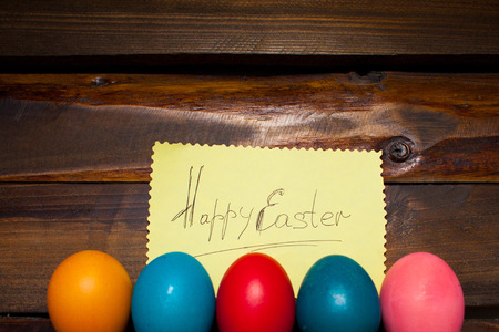 Closed, painted in different bright colors chicken eggs for the holiday Easter on natural wooden background with sheet of paper and the inscription Happy Easter.の写真素材