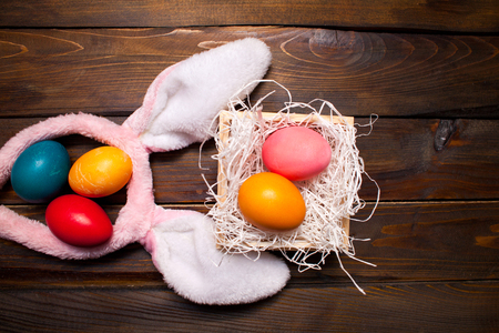 Multicolored chicken, painted eggs for the Easter holiday in a box on a wooden out-looking background with pink rabbit ears.の写真素材