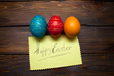 Closed, painted in different bright colors chicken eggs for the holiday Easter on natural wooden background with sheet of paper and the inscription Happy Easter.の写真素材