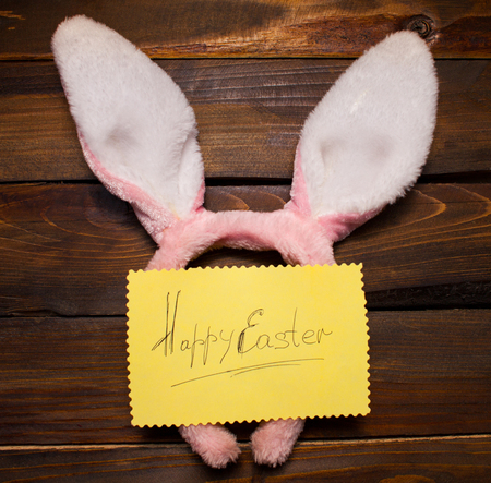 Pink rabbit ears and yellow piece of paper with the inscription Happy Easter on natural, wooden background.の写真素材