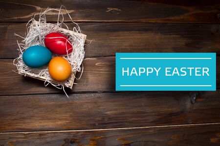 Multi-colored, bright Easter eggs in box with tinsel on wooden background with white text Happy Easter on blue rectangle.の写真素材