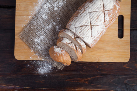 Fresh brown bread with sliced pieces on an oak board with scattered flour on a wooden background with a piece of linen fabric. Photo top view.の写真素材