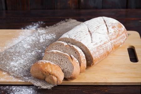Fresh brown bread with sliced pieces on an oak board with scattered flour on a wooden background with a piece of linen fabric.の写真素材