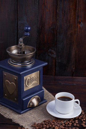 White cup of coffee next to a coffee grinder and ripe coffee grains on a dark wooden background with linen cloth. Vertical photo.の写真素材