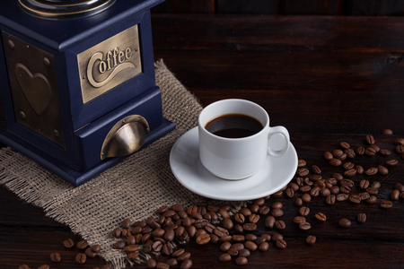 White cup of coffee next to a coffee grinder and ripe coffee grains on a dark wooden background with linen cloth.の写真素材