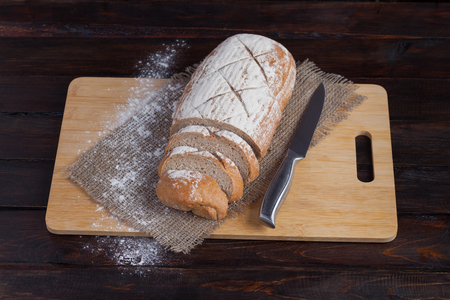 Fresh brown bread with sliced pieces on an oak board with scattered flour on a wooden background with of linen fabric with a steel knife. Photo top viewの写真素材
