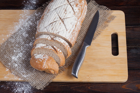 Fresh brown bread with sliced pieces on an oak board with scattered flour on a wooden background with of linen fabric with a steel knife. Photo top viewの写真素材