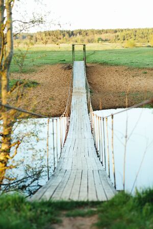 Old suspended wooden foot bridge over the river. Vertical photo on a summer day. River crossing for peopleの写真素材