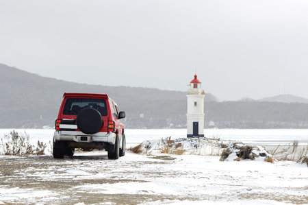 Red car in front of lighthouse on the seacoast in snowy winter dayの写真素材