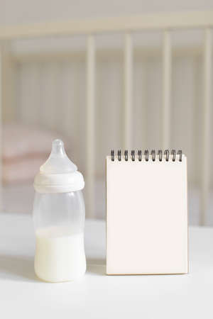 Bottle with milk for baby and empty paper notepad in the background of baby bed. Newborn baby and child care concept. Free copy space.の写真素材