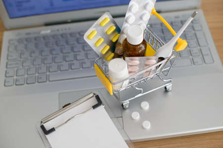 Shopping cart toy with medicaments and paper note in front of laptop screen with pharmacy web site on it. Pills, blister packs, medical bottles, thermometer and recipe set. Health care and internet shopping. Top view.の写真素材