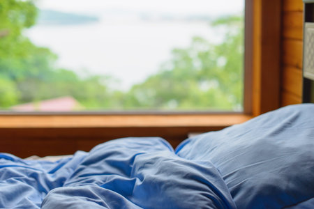 Bed with blue linen in front of the window with blurred nature view in the morning. Sleep with nature concept.の写真素材
