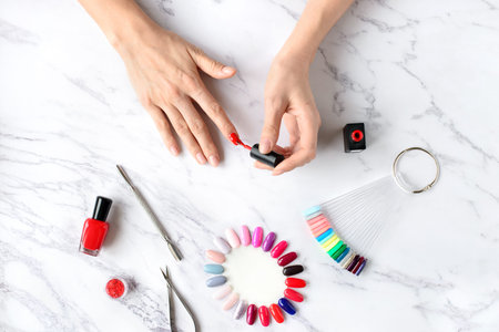 Beautiful woman hands painting nails with red nail polish on marble table with manicure set on it, top view.の写真素材