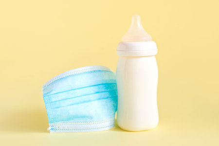 Bottle with breast milk for baby and medical mask on yellow background. Maternity and baby care concept.の写真素材