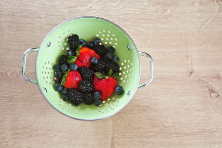 Strawberries, blueberries and blackberries in a colander on a natural wood surfaceの写真素材