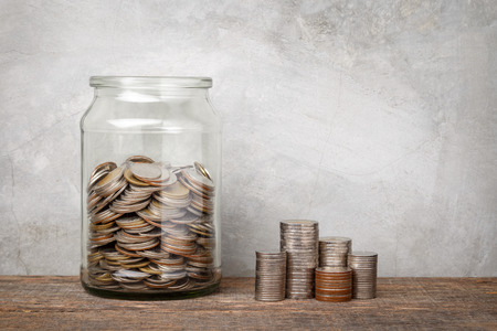 Glass jar with money coins and coins stacked on wooden plank , grey concrete background, money saving financial concept.の写真素材