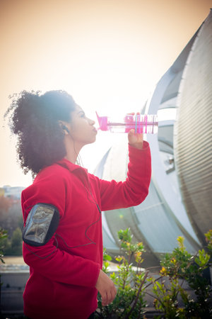 Beautiful athlete woman drinking after running with earphones, smart watch and mobile app in the parkの写真素材