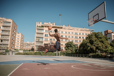 One afroamerican young man without tshirt is playing basketball in a park in Madrid during summer at midday. He is doing an awesome jump to make a dunk and put the ball into the basket.の写真素材