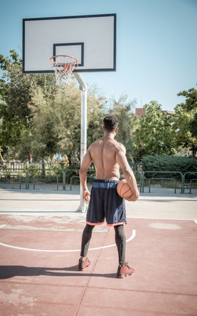 One afroamerican young man without tshirt is playing basketball in a park in Madrid during summer at...