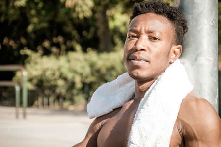 One afroamerican young man without tshirt is playing basketball in a park in Madrid during summer at midday. He is seated on the court while drinking water and cleaning his sweat with a white towel. The ball is under his knees. He is resting after doing excercise.の写真素材