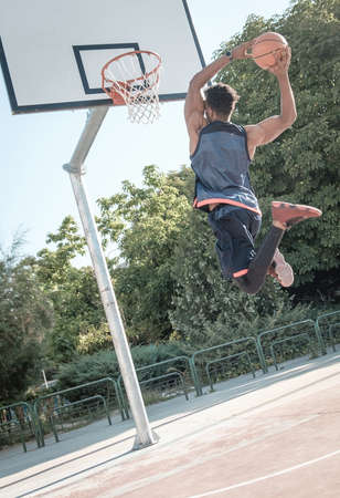 One afroamerican young man is playing basketball in a park in Madrid during summer at midday. He is doing an awesome jump to make a dunk and put the ball into the basket.の写真素材