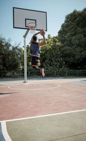 One afroamerican young man is playing basketball in a park in Madrid during summer at midday. He is doing an awesome jump to make a dunk and put the ball into the basket.の写真素材