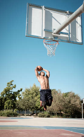 One afroamerican young man without tshirt is playing basketball in a park in Madrid during summer at midday. He is doing an awesome jump to make a dunk and put the ball into the basket.の写真素材