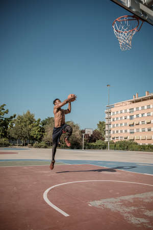 One afroamerican young man without tshirt is playing basketball in a park in Madrid during summer at midday. He is doing an awesome jump to make a dunk and put the ball into the basket.の写真素材