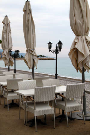 Restaurant tables, chairs and folded umbrellas at seaside on cloudy dayの写真素材