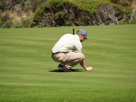 A golfer aligns his putt on the green, holding his ball and a putter.の写真素材