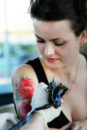 tattooer showing process of making a tattoo on young beautiful hipster woman with red curly hair. Tattoo design in the form of rose. Vintage style pictureの写真素材