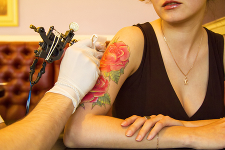 tattooer showing process of making a tattoo on young beautiful hipster woman with red curly hair. Tattoo design in the form of rose. Vintage style pictureの写真素材