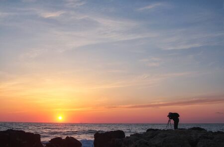 Silhouette of a photographer with a backpack and tripod with a beautiful sea sunset on the backgroundの写真素材