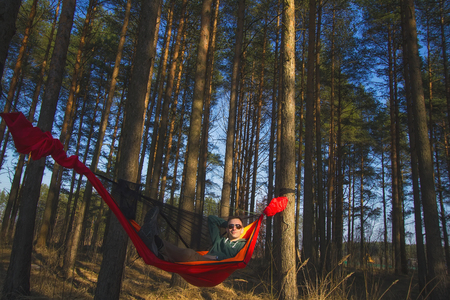 Tourist with sunglasses resting in a red hammock in a pine forest at spring sunset. Travel and adventureの写真素材