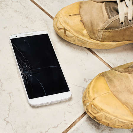 Smart phone with the broken screen over the ceramic floor tiles next to a pair of dirty working shoesの写真素材