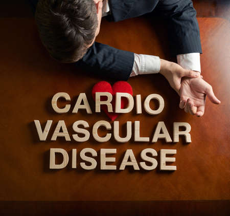 Phrase Cardio Vascular Disease made of wooden block letters and devastated middle aged caucasian man in a black suit sitting at the table with the red symbolic heart, top view composition with dramatic lightingの写真素材
