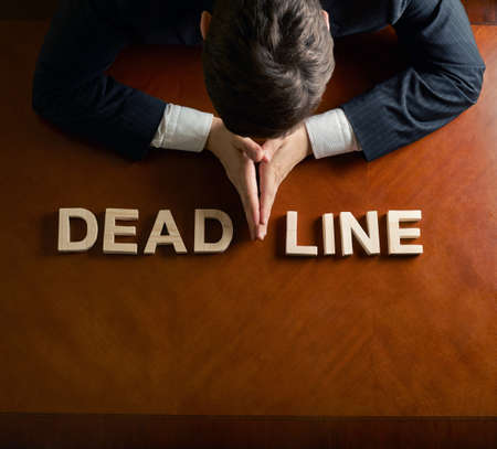 Word Deadline made of wooden block letters and devastated middle aged caucasian man in a black suit sitting at the table, top view composition with dramatic lightingの写真素材