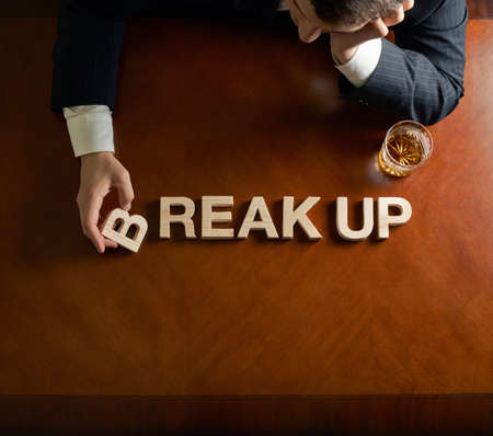 Words Break Up made of wooden block letters and devastated middle aged caucasian man in a black suit sitting at the table with the glass of whiskey, top view composition with dramatic lightingの写真素材