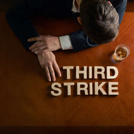 Phrase Third Strike made of wooden block letters and devastated middle aged caucasian man in a black suit sitting at the table with the glass of whiskey, top view composition with dramatic lightingの写真素材