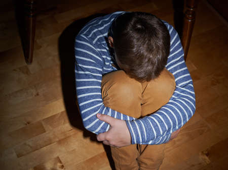 Child abuse composition of a frightened young boy sitting on the wooden floor in a light of a flashlight circleの写真素材