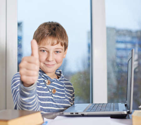 Happy 12 years old children boy sitting at the wooden desk against the notebook computer while showing a thumbs up gesture, composition against the windowの写真素材