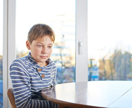 Bemused 12 years old children boy sitting at the wooden desk, composition against the windowの写真素材