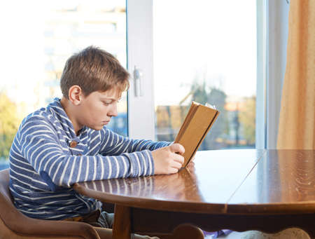 12 years old children boy sitting at the wooden desk while studying and reading a book, composition against the windowの写真素材
